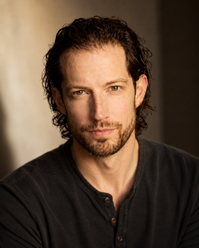 Tim, looking directly at the camera against a warmly lit textured wall, dark henley